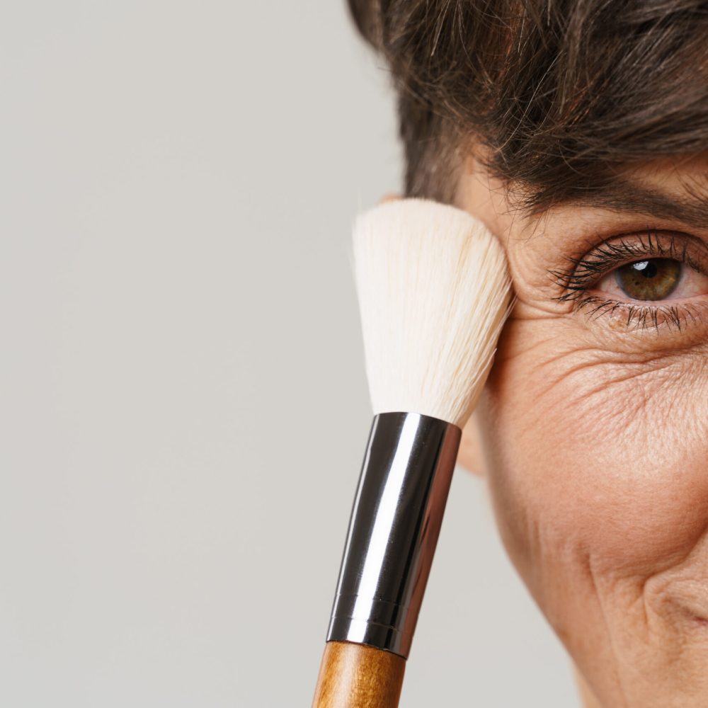 Mature half-naked woman smiling while using powder brush isolated over white background